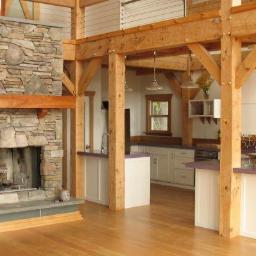 Bud-Ma - Wnętrze domu szkieletowego z kamiennym kominkiem, drewnianą konstrukcją nośną, jasną kuchnią i drewnianą podłogą.