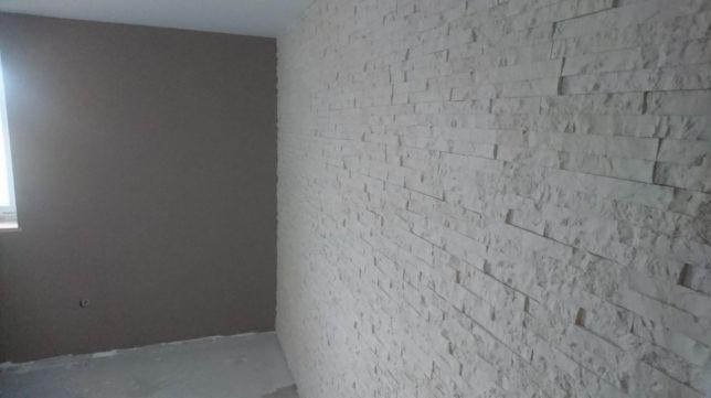 Pokój po remoncie: ściana z dekoracyjnej, białej cegły, druga ściana pomalowana na szaro, widoczne okno i surowa podłoga.