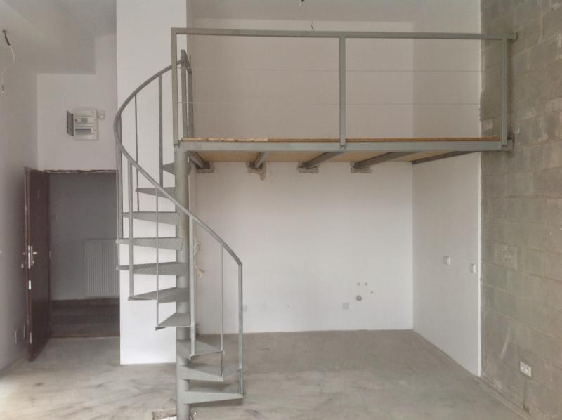 Wnętrze w trakcie remontu z metalowymi schodami spiralnymi prowadzącymi na antresolę z drewnianą podłogą, surowe ściany i betonowa posadzka.