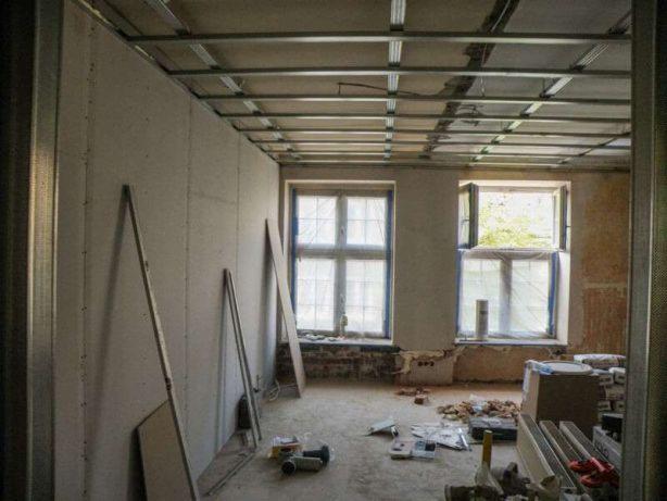 Wnętrze w trakcie remontu: metalowa konstrukcja sufitu podwieszanego, częściowo obłożone płyty gipsowo-kartonowe na ścianach, okna z zasłoniętymi szybami, narzędzia i materiały budowlane na podłodze.