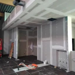 Wnętrze w trakcie prac wykończeniowych: widoczne ściany i sufit z płyt gipsowo-kartonowych, częściowo wyszpachlowane, przewody elektryczne wystające z podłogi, drabina oparta o ścianę, wentylacja...