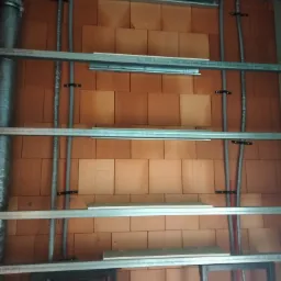 Ceglana ściana z zamontowaną metalową konstrukcją pod przyszłą okładzinę, widoczne przewody elektryczne i rury przymocowane do ściany