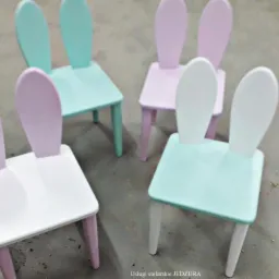 Cztery dziecięce krzesła w pastelowych kolorach (miętowy, lawendowy i biały) z oparciami w kształcie króliczych uszu, ustawione na szarej podłodze.