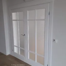 Białe, dwuskrzydłowe drzwi drewniane z mlecznym szkłem w prostokątnych kasetonach, widoczne na tle parkietu i białych ścian z gniazdkami.