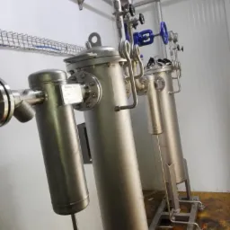 Filtracja przemysłowa: zestaw trzech cylindrycznych filtrów ze stali nierdzewnej z manometrami i zaworami, zamontowanych na metalowych podstawach w przemysłowym pomieszczeniu z panelami ściennymi.