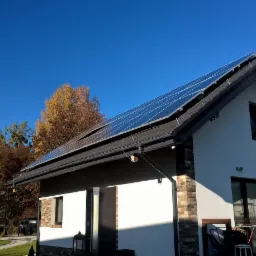 Dom jednorodzinny z panelami słonecznymi na dachu, widoczne okna, drzwi przesuwne i antena satelitarna, jesienne drzewa w tle.