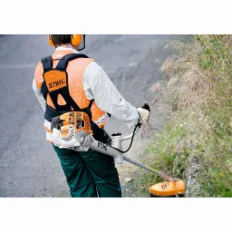 Operator w pomarańczowej kamizelce Stihl i słuchawkach, obsługujący wykaszarkę spalinową Stihl FS 310 przy krawędzi drogi, z widocznymi wysokimi trawami i chwastami.
