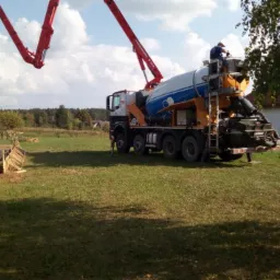 Betonomieszarka z pompą podaje beton do szalunku fundament&oacute;w domu na działce z zieloną trawą, widoczny pracownik obsługujący pompę i częściowo zarys samochodu w prawym dolnym rogu.