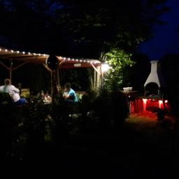 Systemy Elektryczne - Wieczorne oświetlenie altany ogrodowej z grillem, ozdobione lampkami i kolorowymi światłami LED, w tle ciemne drzewa i budynek.