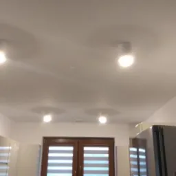Biały sufit z czterema okrągłymi lampami punktowymi LED, widoczne fragmenty zabudowy kuchennej i okno z roletami w tle.