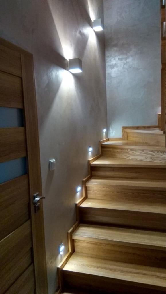 Drewniane schody z oświetleniem LED wbudowanym w ścianę i stopnie, ściana wykończona dekoracyjnym tynkiem, widoczna część drewnianych drzwi.