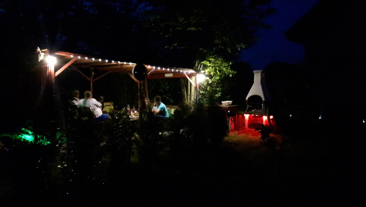 Wieczorne oświetlenie altany ogrodowej z grillem, ozdobione lampkami i kolorowymi światłami LED, w tle ciemne drzewa i budynek.