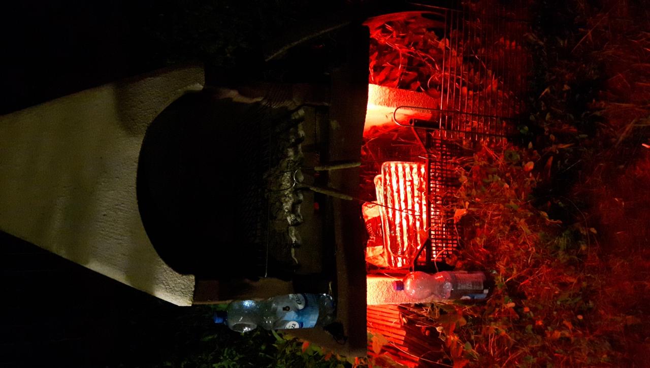 Nocne oświetlenie grilla z czerwonym światłem, z widocznymi butelkami wody i elementami konstrukcyjnymi grilla.