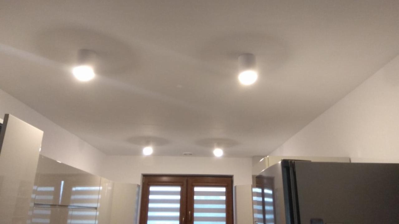 Biały sufit z czterema okrągłymi lampami punktowymi LED, widoczne fragmenty zabudowy kuchennej i okno z roletami w tle.