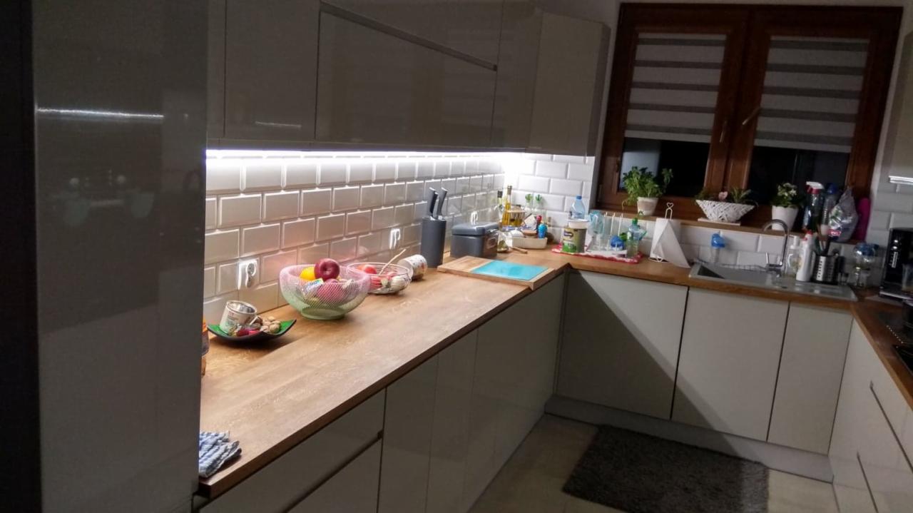 Nowoczesna kuchnia z oświetleniem LED podszafkowym, białymi płytkami typu metro i drewnianym blatem, widok na okno z roletami poziomymi.