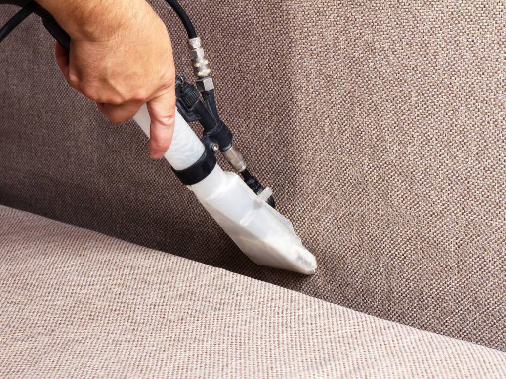 Proces czyszczenia jasnobrązowej sofy za pomocą odkurzacza piorącego z przezroczystą końcówką, widoczna struktura materiału.