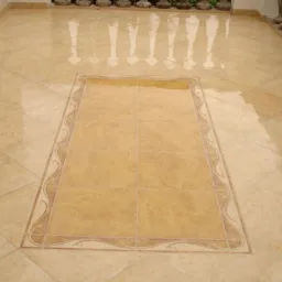 Beżowa podłoga z płytek ceramicznych ułożonych w karo, z centralnym prostokątnym wzorem obramowanym dekoracyjnym pasem.