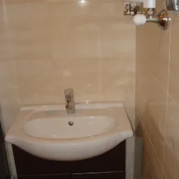 Umywalka nablatowa z brązową szafką, chromowaną baterią i grzejnikiem drabinkowym na beżowych, błyszczących płytkach w łazience po remoncie.