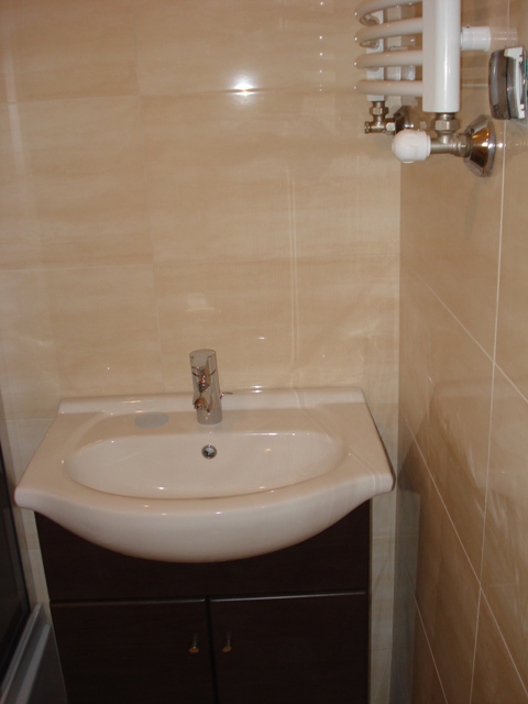 Umywalka nablatowa z brązową szafką, chromowaną baterią i grzejnikiem drabinkowym na beżowych, błyszczących płytkach w łazience po remoncie.