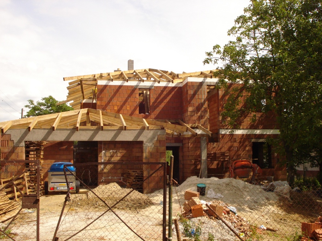 Budowa domu z czerwonej cegły, widoczne drewniane elementy konstrukcyjne dachu, przyczepka samochodowa z boku budynku, teren budowy z piaskiem i cegłami.