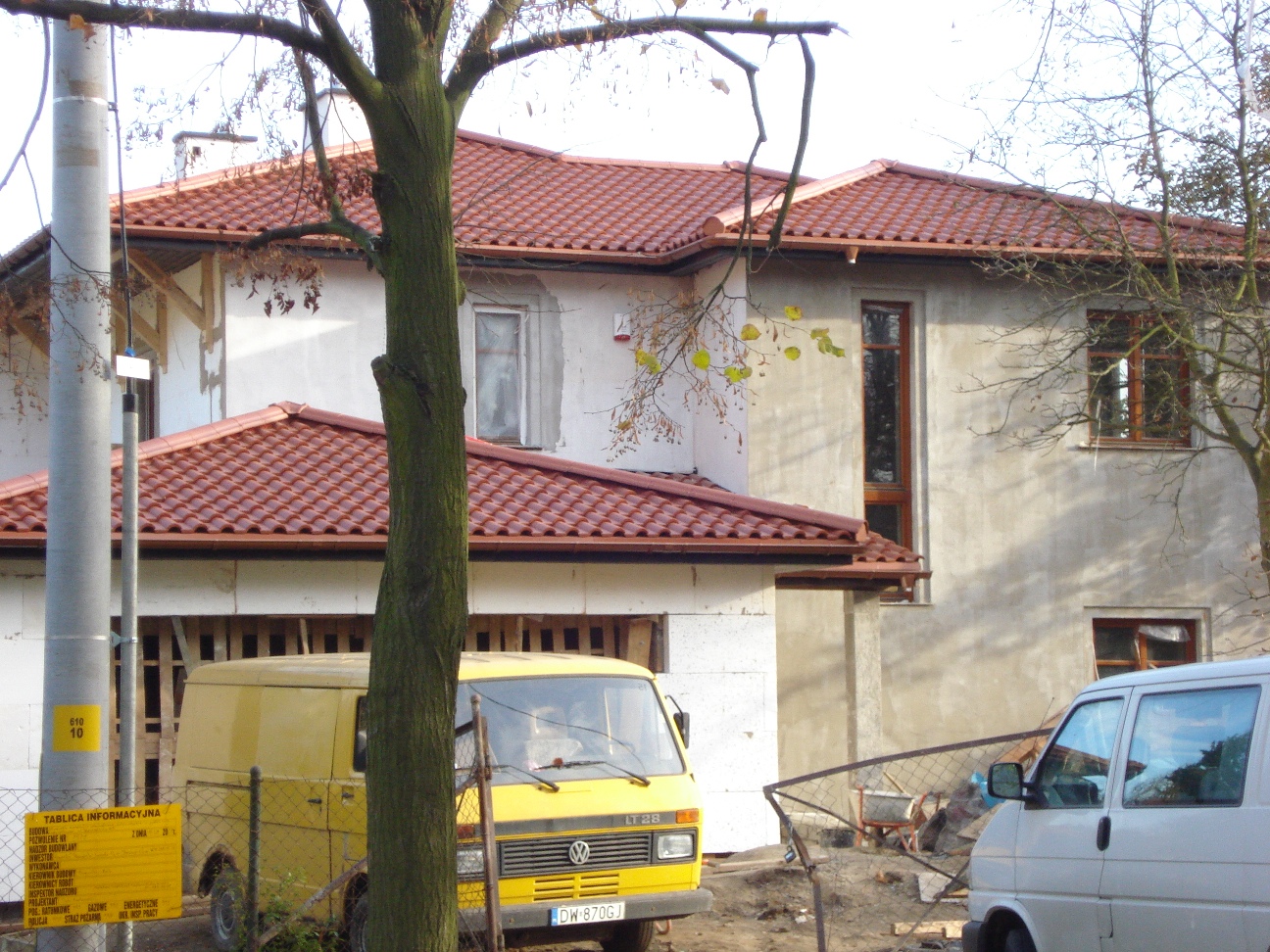 Dom murowany w trakcie budowy z widocznym żółtym samochodem dostawczym i tablicą informacyjną na pierwszym planie, z częściowo wykończonym dachem z czerwonej dachówki.