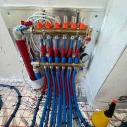 Rozdzielacz ogrzewania podłogowego z czerwono-niebieskimi rurami, zamontowany na ścianie z widocznym manometrem i odpowietrznikiem, ułożony na srebrnej folii izolacyjnej z czerwoną siatką, z żółtą...