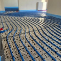 PHU BioTech Łukasz Wańkowicz - Niebieskie rury ogrzewania podłogowego ułożone na folii z siatką, mocowane czarnymi uchwytami. Widok z góry, perspektywa zbieżna, jasne wnętrze w tle.