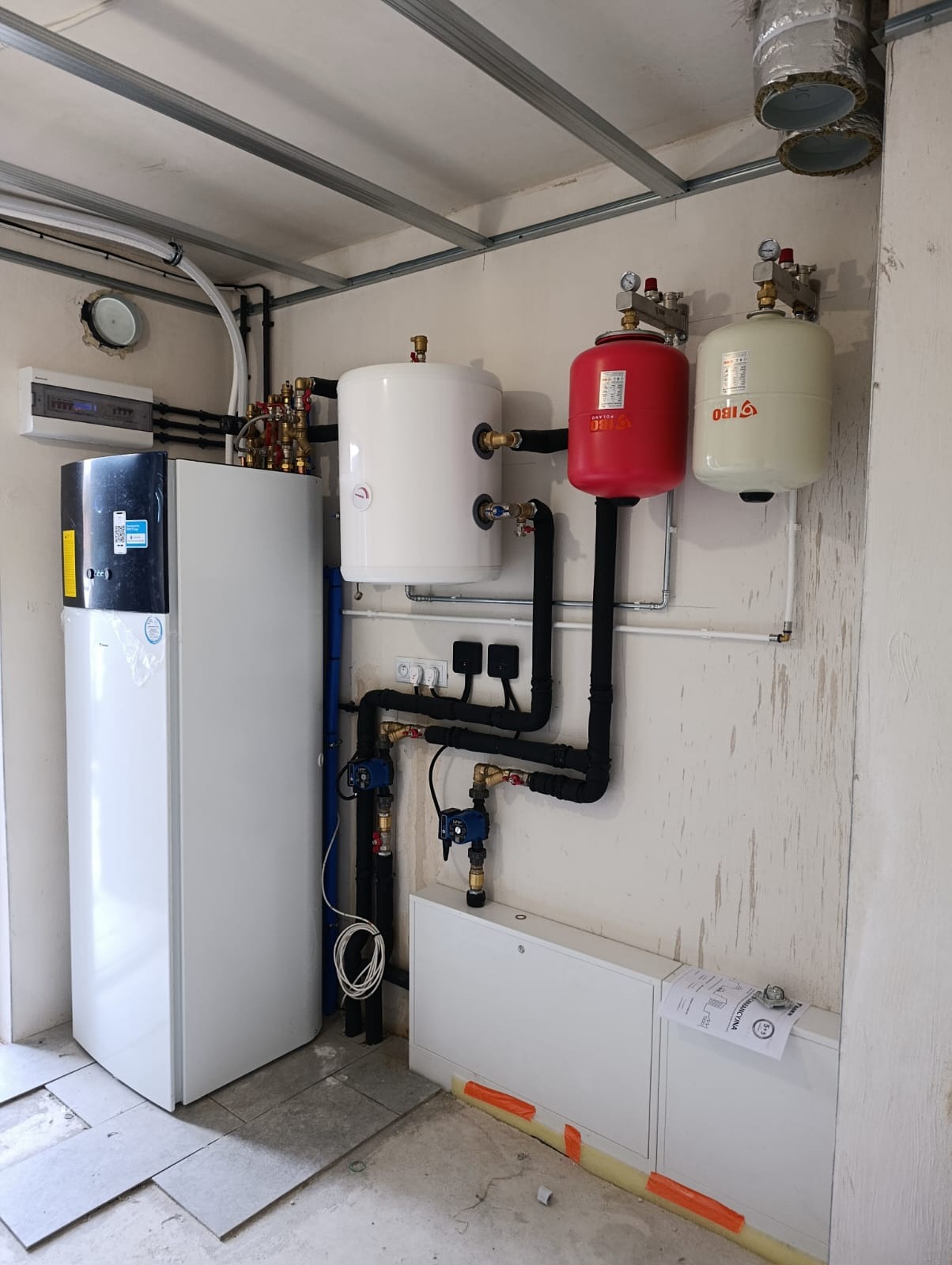 Złożona instalacja grzewcza z białym zbiornikiem, czerwonym naczyniem przeponowym i pompą ciepła w pomieszczeniu z surowym wykończeniem. Widoczne rury i zawory.