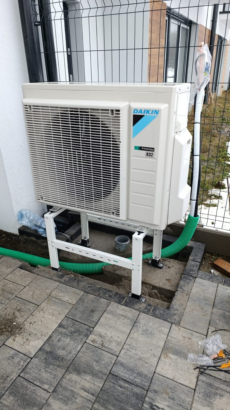 Biała zewnętrzna jednostka klimatyzacji Daikin na metalowym stelażu, z widocznymi zielonymi rurami instalacji, na tle kostki brukowej i ogrodzenia.