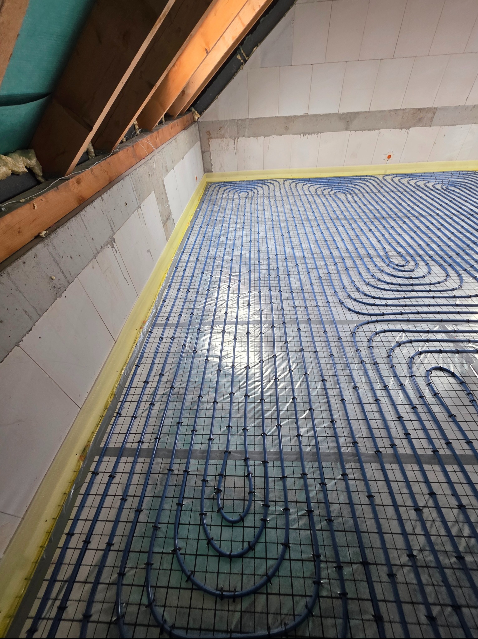 Niebieskie rury ogrzewania podłogowego ułożone na siatce w surowym wnętrzu poddasza, widok z góry, przed wylaniem posadzki. Widoczne drewniane elementy konstrukcyjne dachu.