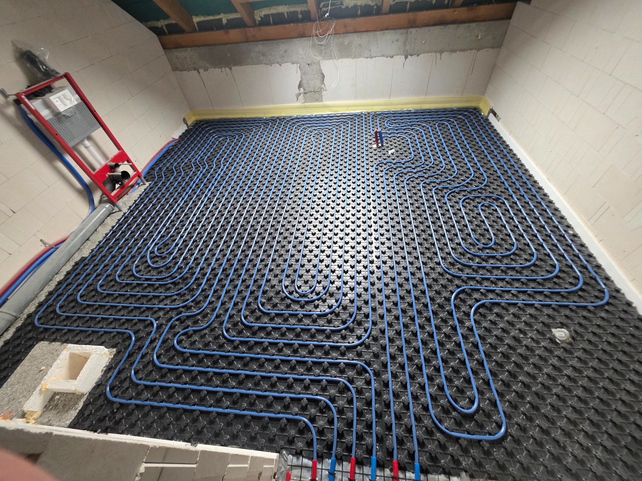 Niebieskie rury ogrzewania podłogowego ułożone na czarnej macie w surowym wnętrzu. Widoczna instalacja sanitarna przy ścianie. Ujęcie z góry, szeroki kadr.