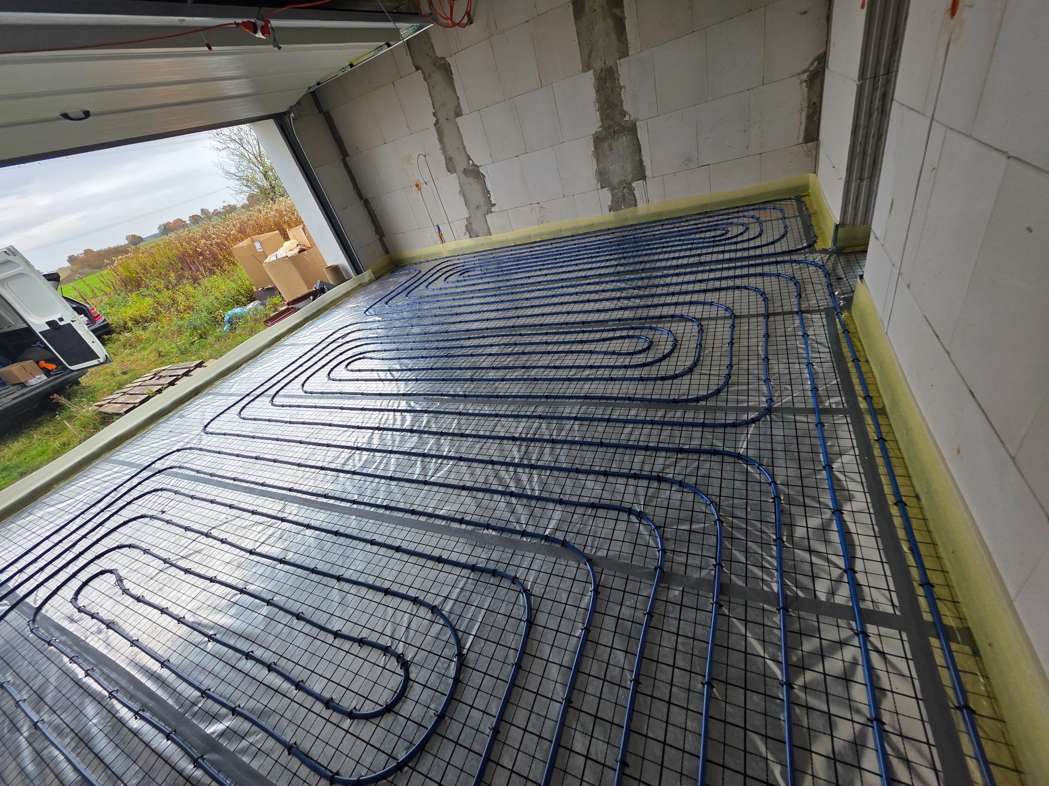 Niebieskie rury ogrzewania podłogowego ułożone na folii w surowym wnętrzu garażu z widokiem na zewnątrz. Widoczny samochód dostawczy i materiały budowlane.