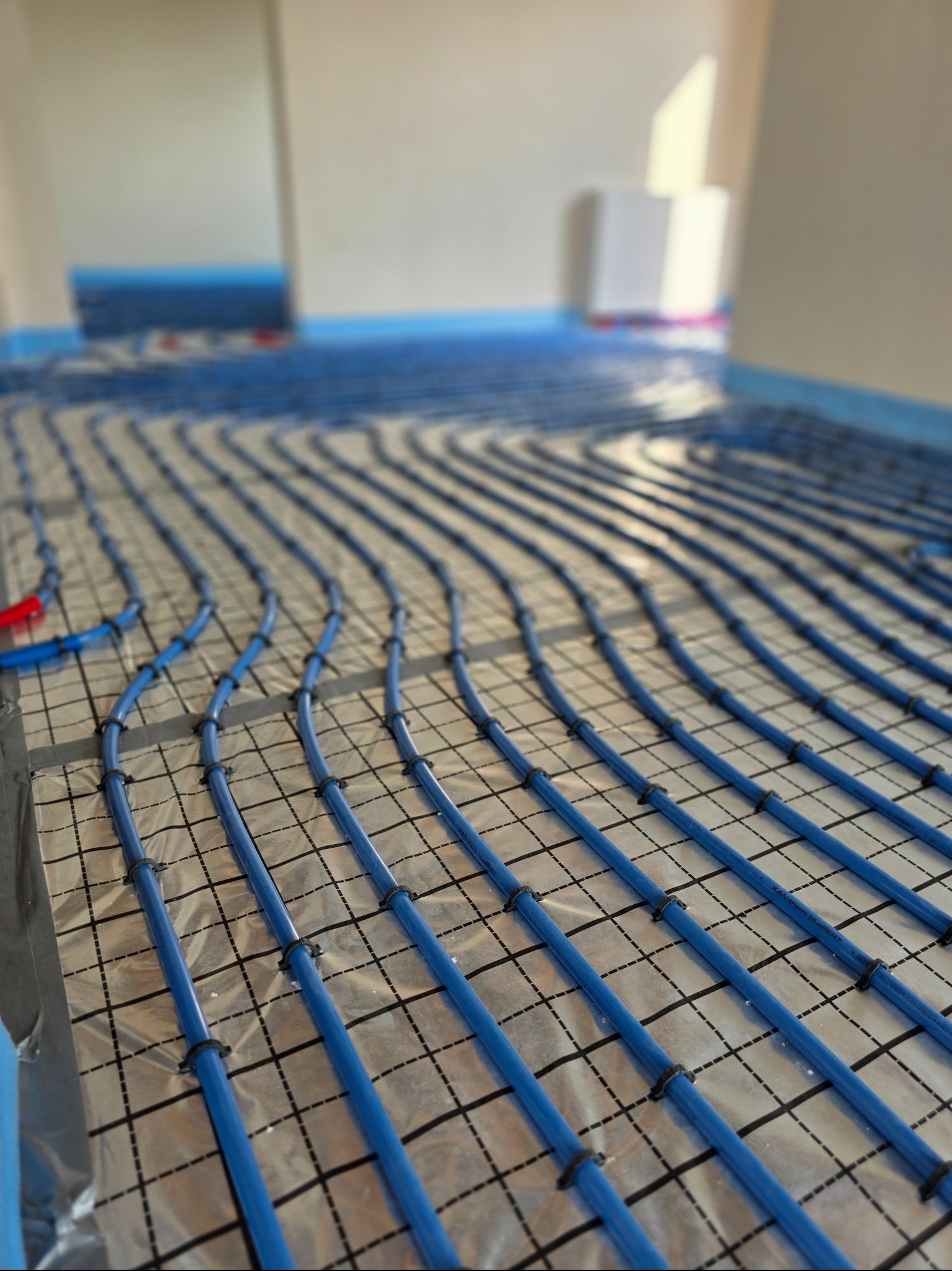 Niebieskie rury ogrzewania podłogowego ułożone na folii z siatką, mocowane czarnymi uchwytami. Widok z góry, perspektywa zbieżna, jasne wnętrze w tle.