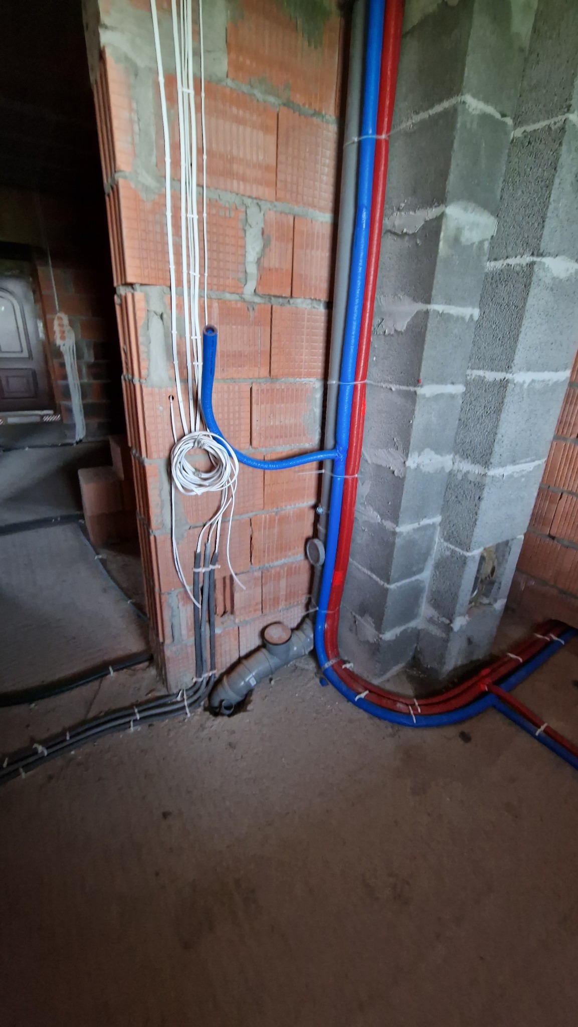 Instalacja wodna z rur w kolorach czerwonym i niebieskim, prowadzone wzdłuż surowej ściany z cegły i betonowego filaru, z widocznymi kablami elektrycznymi i odpływem kanalizacyjnym przy podłodze.