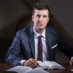 Portret mężczyzny w garniturze siedzącego przy biurku z otwartymi książkami i filiżanką kawy, w tle drewniane elementy wystroju.