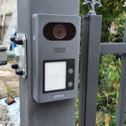 Szary wideodomofon Dahua zamontowany na metalowej bramie z ozdobnymi zakończeniami, widoczne mocowania i elementy konstrukcyjne bramy, w tle zieleń i fragment budynku w trakcie prac.