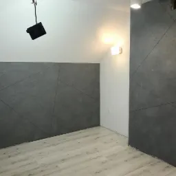 Pokój po remoncie ze ścianą i filarem pokrytymi szarą, dekoracyjną strukturą z geometrycznym wzorem, jasne panele na podłodze, surowe oświetlenie.