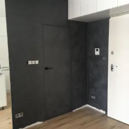 Ściana w odcieniach szarości z ukrytymi drzwiami, wykończona dekoracyjnym tynkiem imitującym beton, widoczne gniazdka elektryczne i drewnopodobna podłoga.