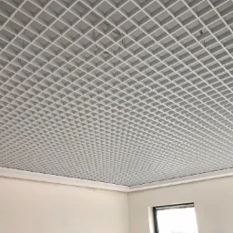 Biały sufit rastrowy z widocznymi elementami mocującymi i fragmentami ściany oraz okna w pomieszczeniu.