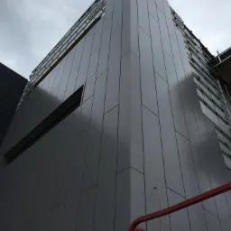 Elewacja budynku w trakcie montażu z wykorzystaniem paneli w odcieniach szarości, widoczne rusztowania i fragment podnośnika. Ujęcie z dołu, pochmurne niebo.