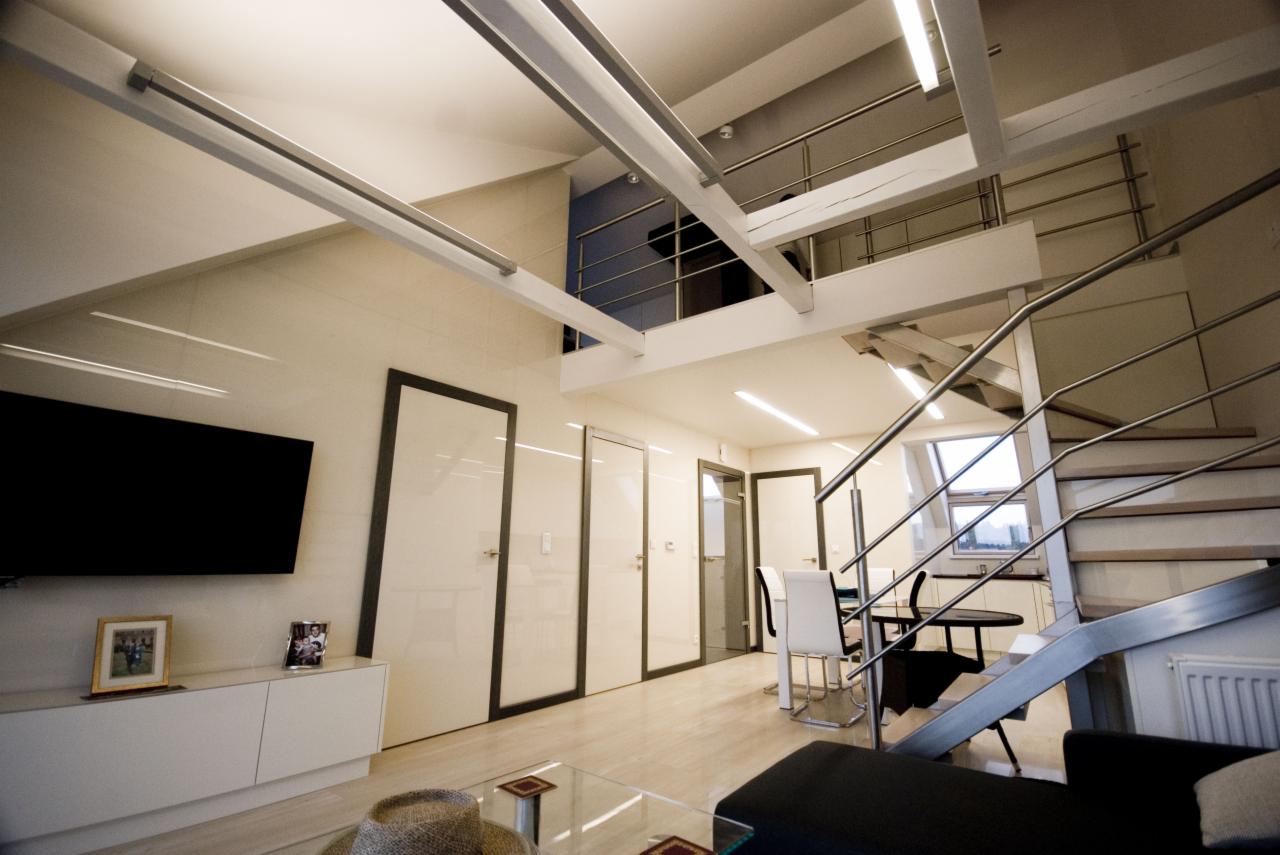 Nowoczesne wnętrze dwupoziomowego mieszkania z antresolą, metalowymi schodami, białymi drzwiami z ciemnymi ramami, telewizorem i minimalistycznymi meblami. Na pierwszym planie szklany stolik kawowy...