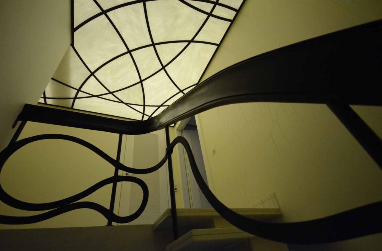 Klatka schodowa z dekoracyjną balustradą z czarnego metalu i podświetlanym sufitem z geometrycznym wzorem, widok z dołu.