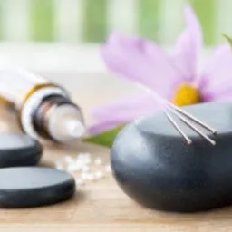 Trzy igły do zabiegów na gładkim, czarnym kamieniu obok buteleczki z kroplomierzem, małych kamyczków i fioletowego kwiatu na drewnianym podłożu.