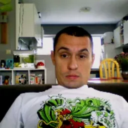 Mężczyzna w białej koszulce z wzorem na tle kuchni z białymi szafkami i żółtym krzesłem. Ujęcie z bliska, widoczna twarz i ramiona.