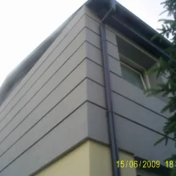 Fragment narożnika budynku z szarą elewacją w pasy, rynną i zielonym drzewem iglastym w tle.