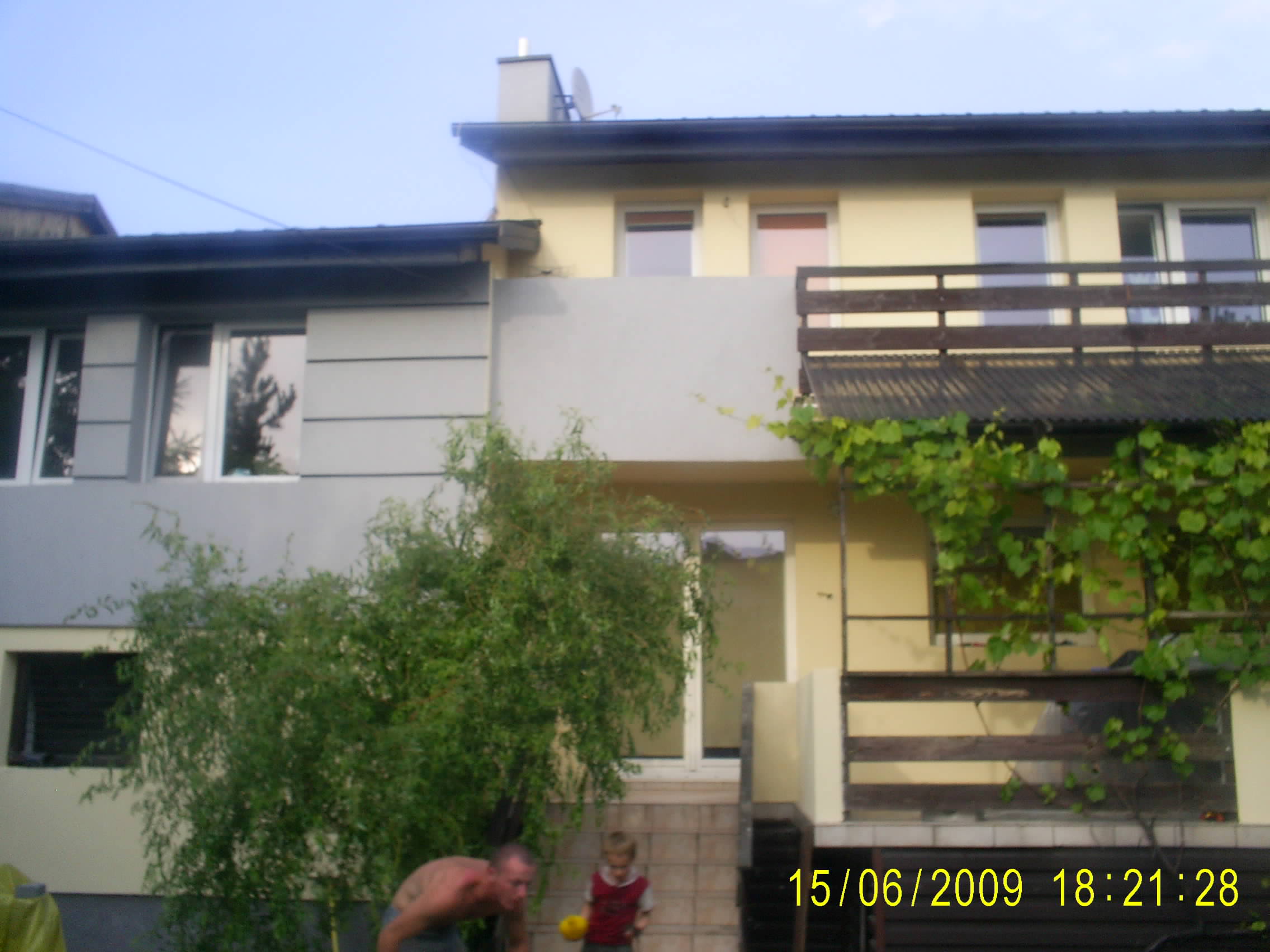 Dwukondygnacyjny dom z częściowo ocieploną elewacją, widoczne fragmenty szarej i żółtej ściany, z przodu rosnąca zieleń, na pierwszym planie mężczyzna i dziecko.