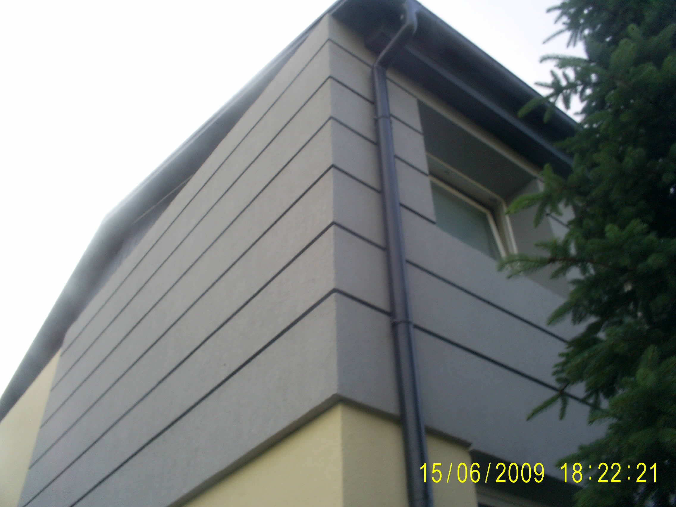 Fragment narożnika budynku z szarą elewacją w pasy, rynną i zielonym drzewem iglastym w tle.