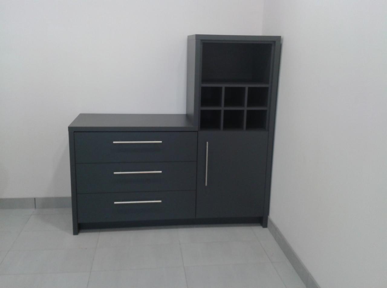 Nowoczesna, grafitowa komoda z trzema szufladami i szafką z półkami na wino, ustawiona w rogu pomieszczenia na szarej podłodze.