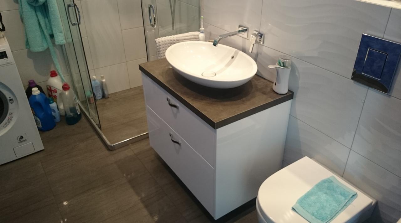 Nowoczesna łazienka z białą szafką pod umywalkę z szufladami, brązowym blatem i owalną umywalką nablatową. W tle kabina prysznicowa, pralka i płytki w odcieniach bieli i szarości.