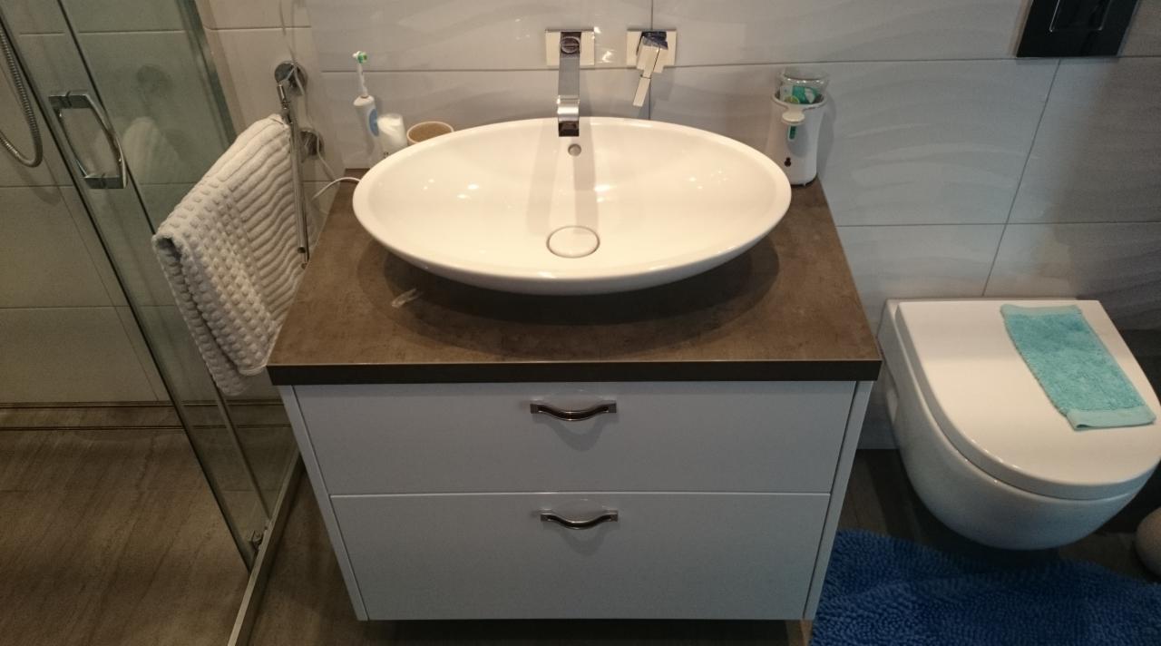 Nowoczesna łazienka z owalną umywalką nablatową na szafce z szufladami, minimalistyczna armatura, płytki w odcieniach szarości i kabina prysznicowa ze szklanymi drzwiami po lewej stronie.